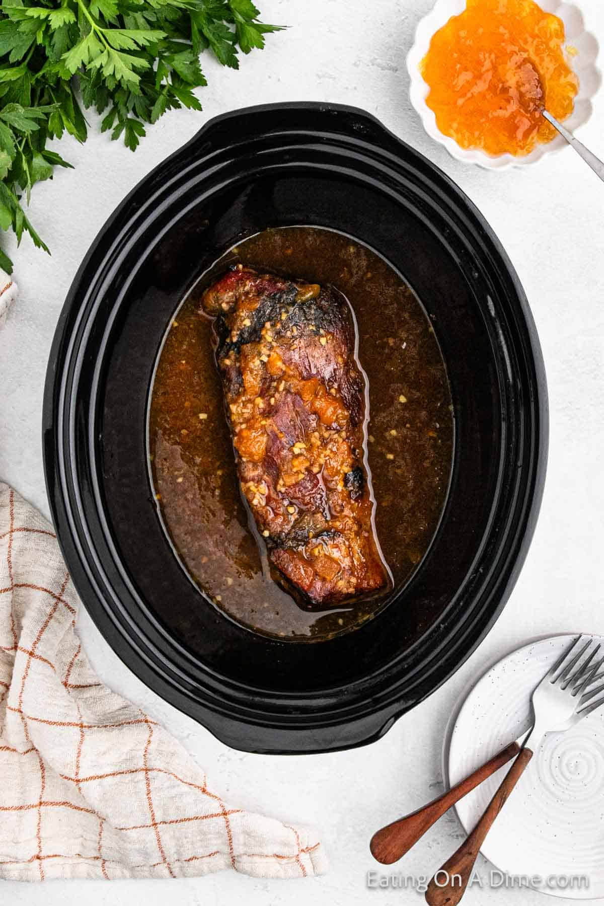 A Crock Pot holds a well-cooked pork tenderloin seasoned with spices and garnished with a glaze. Surrounding the cooker are fresh parsley, a bowl of orange peach sauce, a napkin, and a small plate with wooden utensils.