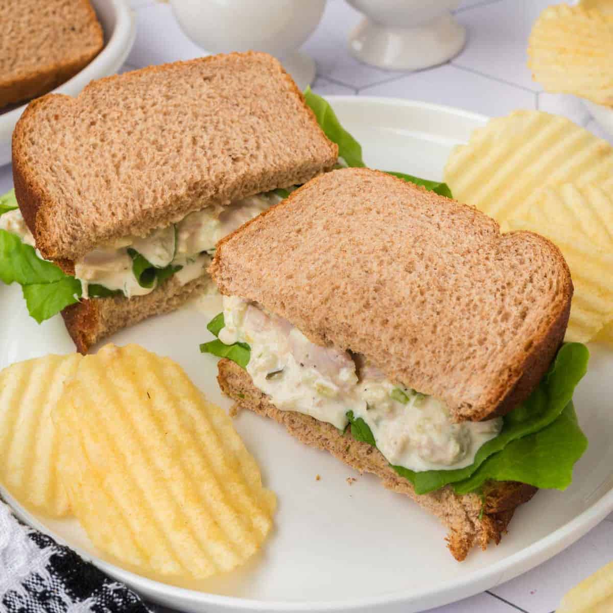 A plate with a chicken salad sandwich on whole wheat bread, cut in half and placed side by side. The sandwich contains lettuce and chicken salad filling. Rippled potato chips are served alongside, reminiscent of a classic tuna salad sandwich recipe. A checkered cloth is partially visible in the bottom left corner.