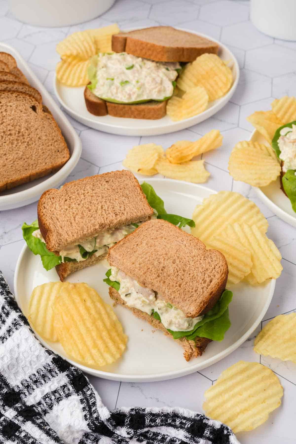 Two plates of chicken salad sandwiches on whole grain bread are served with potato chips. One plate in the foreground includes a black and white checkered napkin, while another plate is in the background, also accompanied by chips. Additional sandwich ingredients, ideal for a tuna salad sandwich recipe, are nearby.