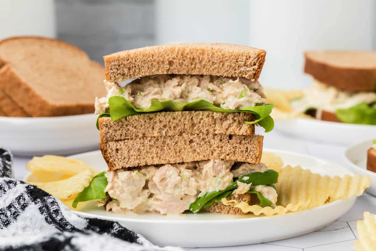 A plate holds a stacked tuna salad sandwich with lettuce on whole wheat bread. Potato chips are on the side, with an additional sandwich and slices of bread in the background. A black-and-white checkered cloth is partially visible in the foreground, suggesting a cozy meal inspired by a favorite tuna salad sandwich recipe.