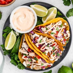 A plate of three easy fish tacos filled with diced tomatoes, red onions, cilantro, and crumbled cheese, topped with a drizzle of creamy sauce. The tacos are accompanied by lime wedges and a bowl of sauce on the side. Fresh cilantro garnishes the plate.