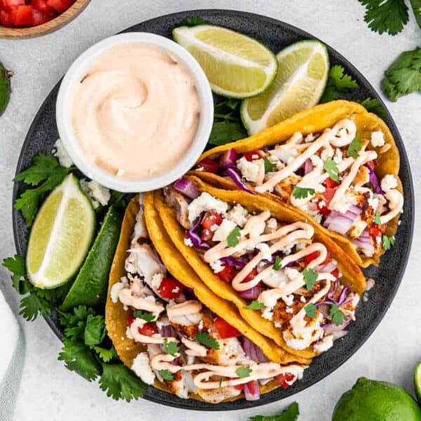 A plate of three easy fish tacos filled with diced tomatoes, red onions, cilantro, and crumbled cheese, topped with a drizzle of creamy sauce. The tacos are accompanied by lime wedges and a bowl of sauce on the side. Fresh cilantro garnishes the plate.