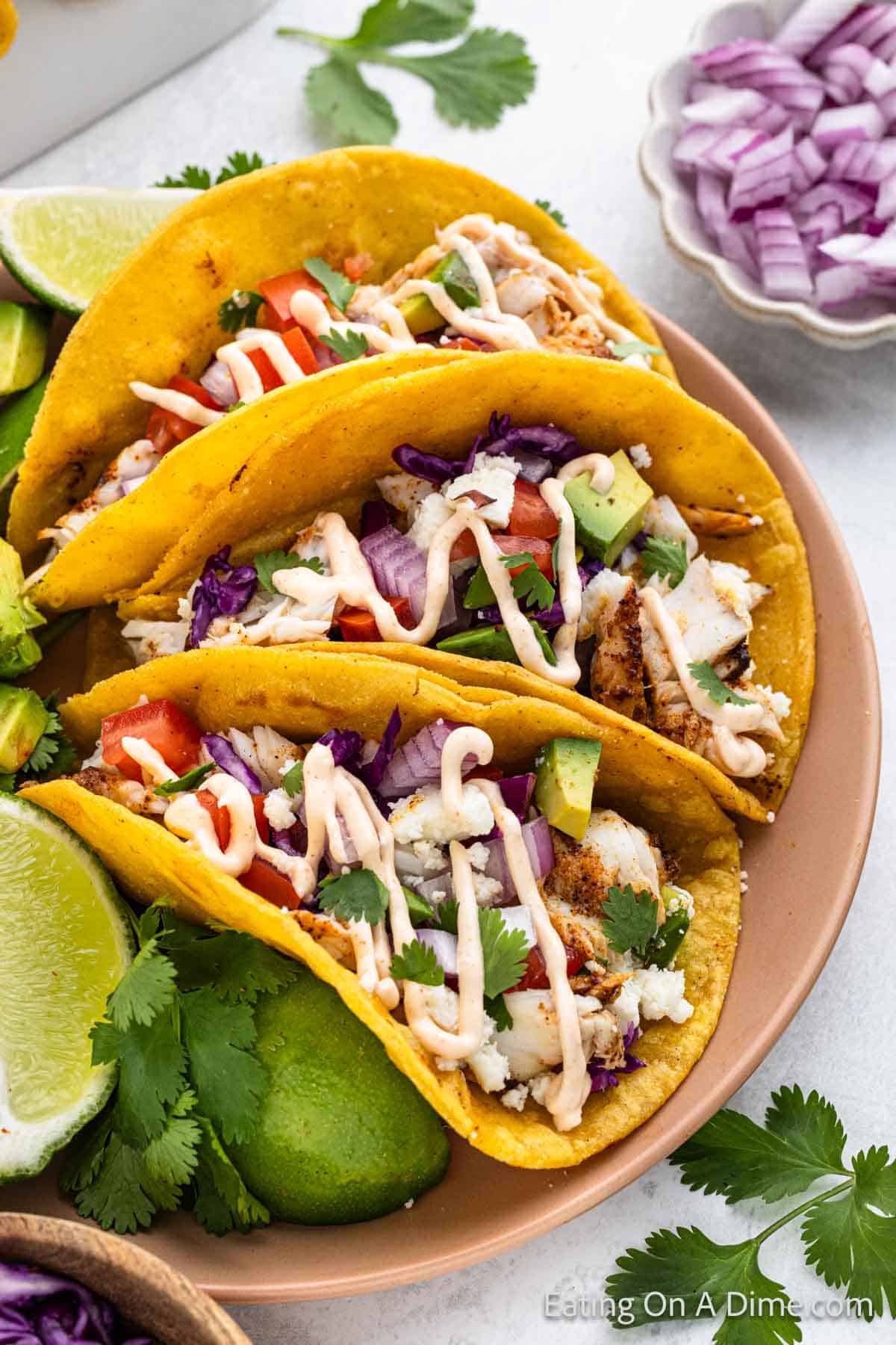 Three fish tacos filled with flaked white fish, shredded red cabbage, diced tomatoes, sliced avocado, cilantro, and topped with a drizzle of creamy sauce. This easy fish tacos recipe is served on a pink plate with lime wedges, avocado halves, and cilantro on the side. Red onion is in a bowl nearby.