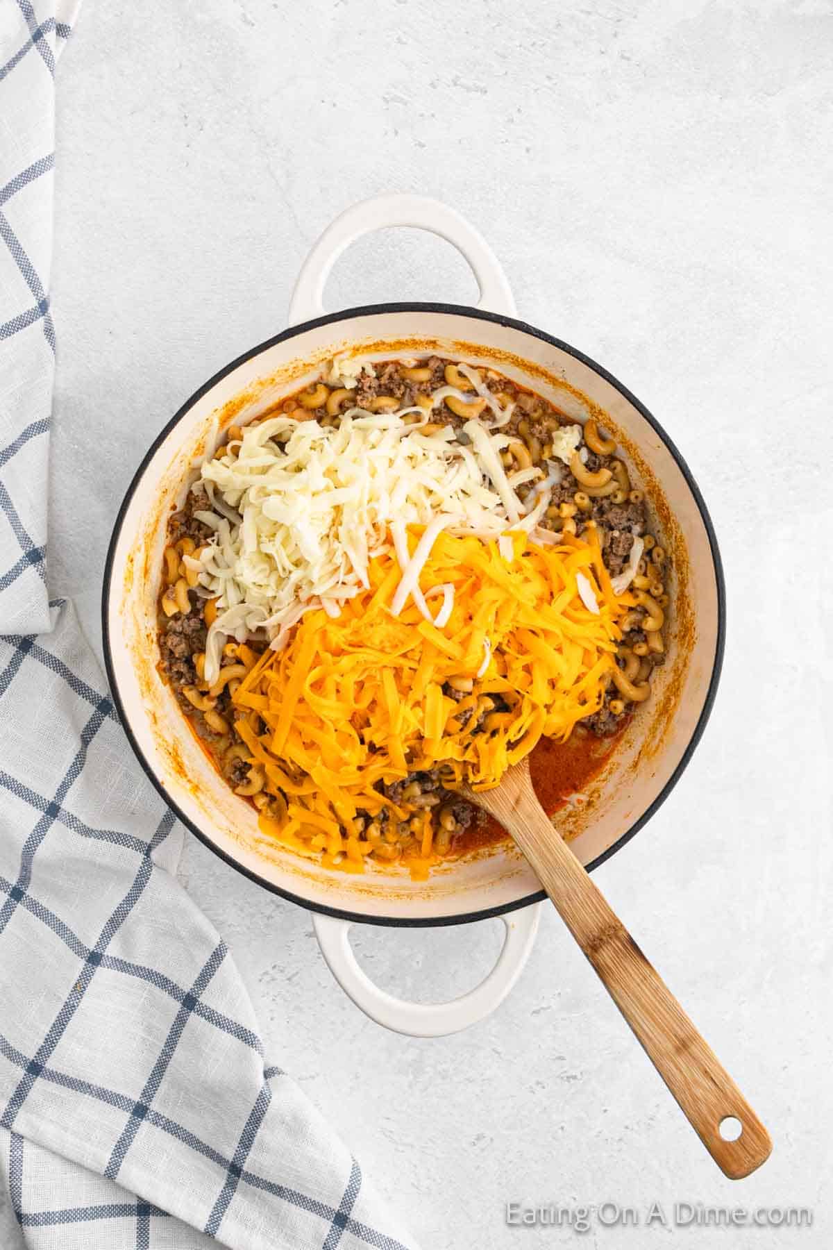 A white pot sits on a white and blue checkered cloth. Inside the pot, there is a mixture of cooked pasta, ground meat, and shredded cheese, with orange and white cheese on top. A wooden spoon rests in the pot, ready to stir this delicious Homemade Cheeseburger Helper together.