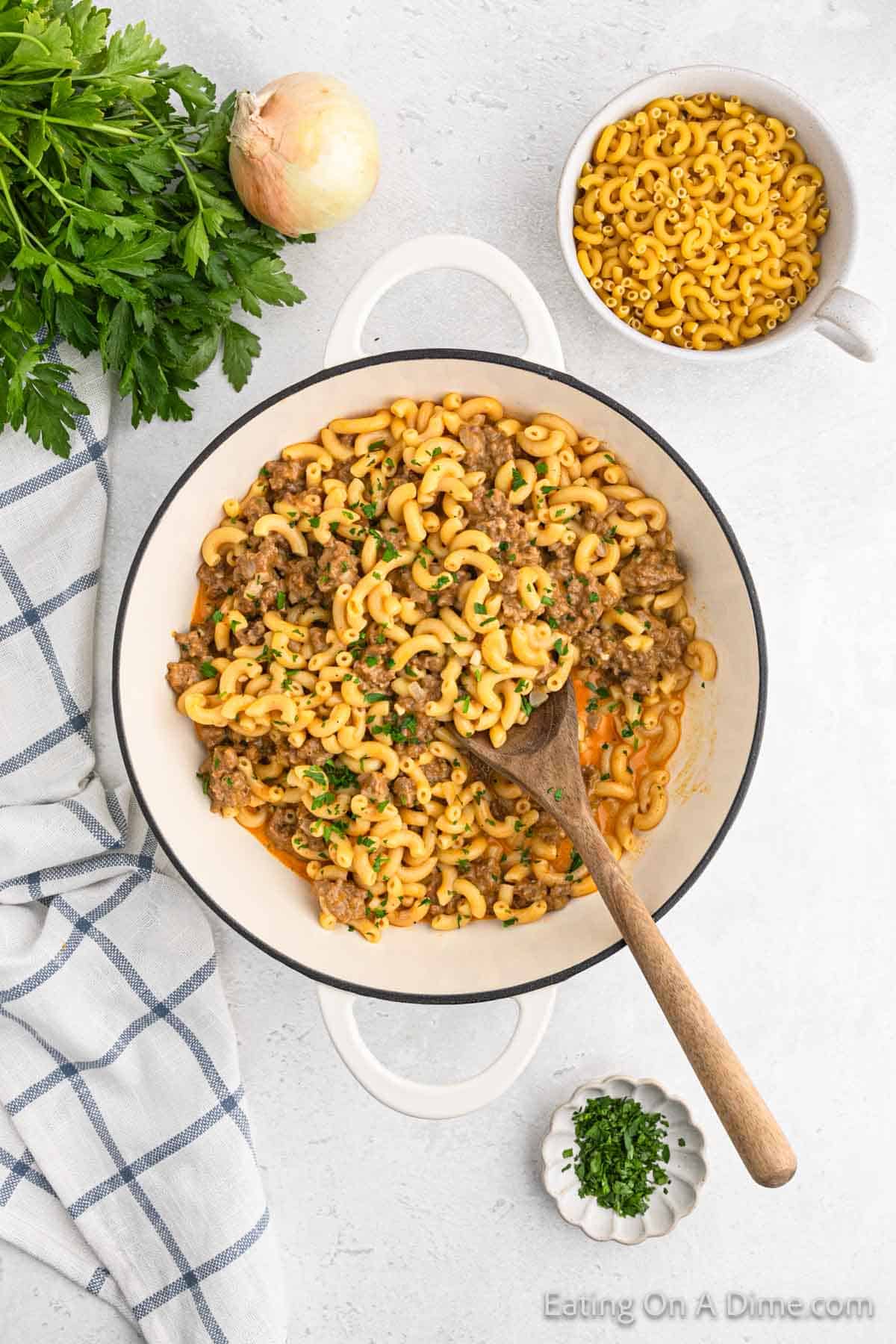 Top view of a skillet filled with Homemade Cheeseburger Helper, featuring macaroni pasta, ground beef, and garnished with chopped parsley. The skillet is surrounded by fresh parsley, an onion, uncooked macaroni, and a blue checkered kitchen towel on a white surface.