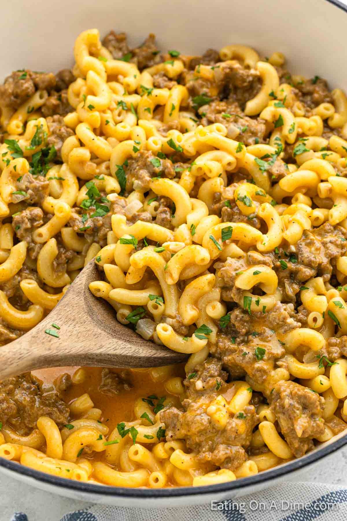 A wooden spoon stirs a pot of creamy homemade Cheeseburger Helper, featuring macaroni mixed with a savory, cheesy sauce and ground beef casserole garnished with chopped parsley for added color and flavor.