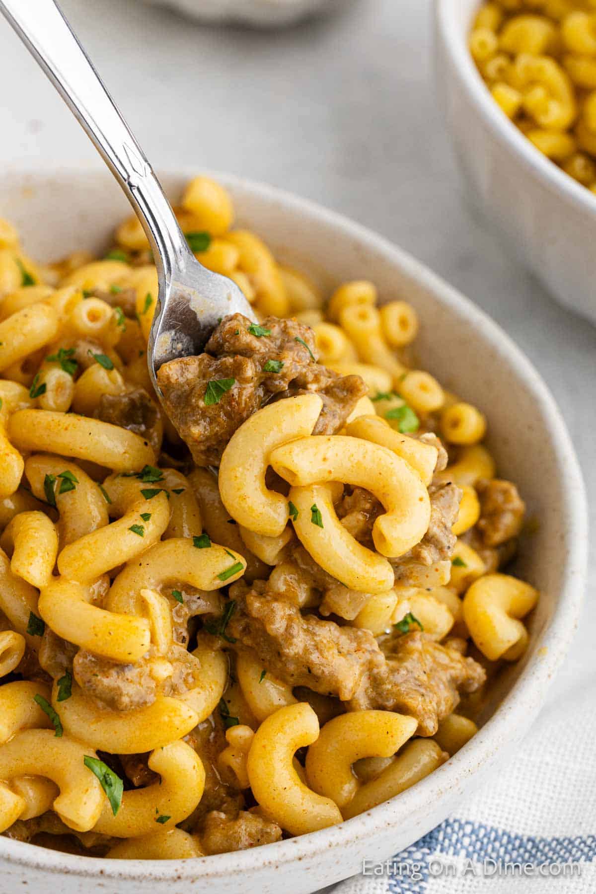 A bowl of creamy macaroni and cheese mixed with ground beef, reminiscent of a homemade Cheeseburger Helper. A fork is lifting a portion of the pasta, showcasing the saucy, savory mixture. The dish is garnished with finely chopped parsley. In the background, a side bowl of corn is visible.