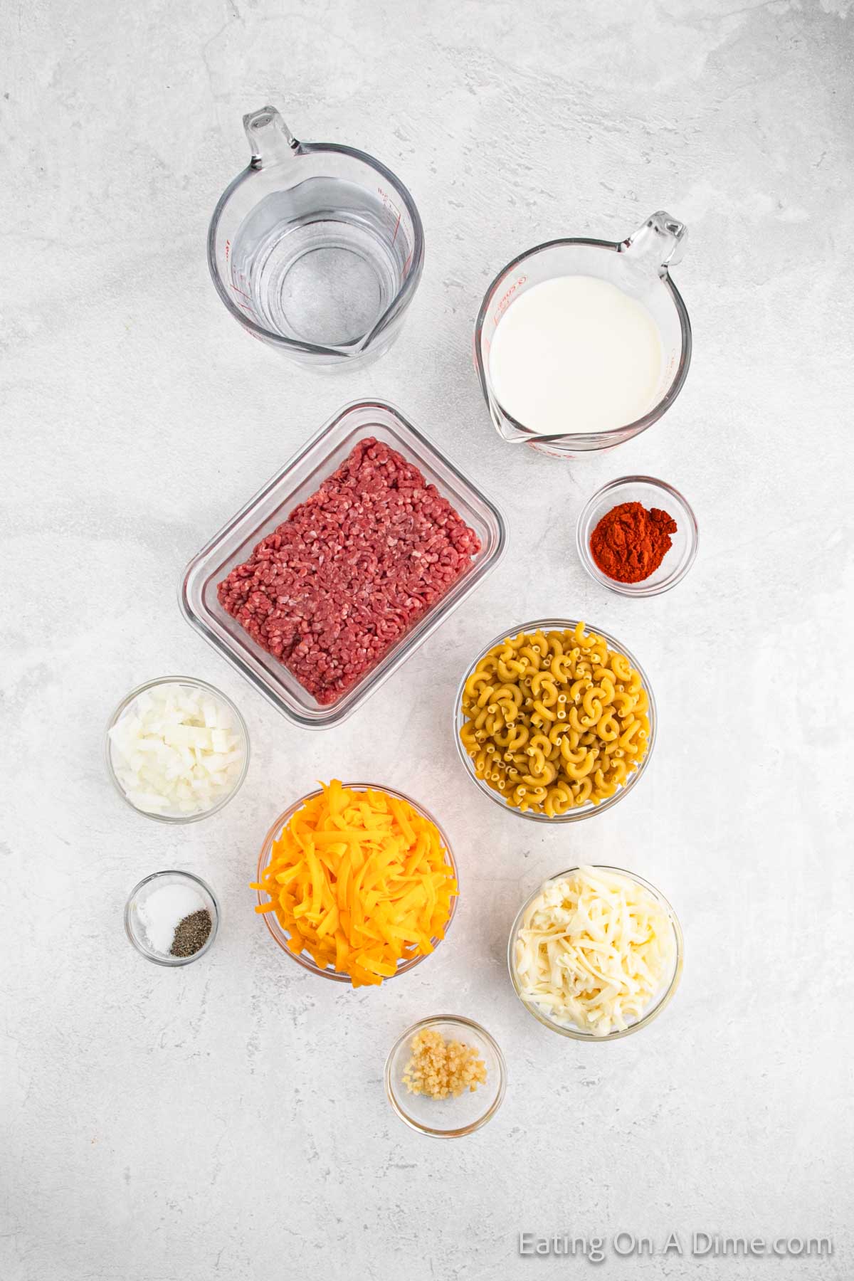 Top view of ingredients for a homemade cheeseburger helper recipe. Various bowls and measuring cups contain elbow macaroni, ground beef, shredded cheese, milk, water, chopped onion, minced garlic, and seasonings, all arranged on a light surface.