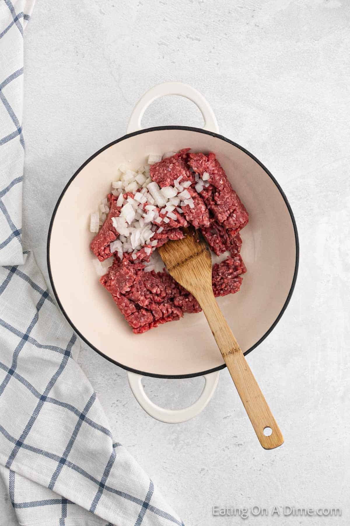 A white pot containing raw ground beef and chopped onions is placed on a light-colored countertop, ready to create a homemade Cheeseburger Helper. A wooden spatula rests inside the pot, while a white and blue checkered cloth is partly visible on the left side of the image.