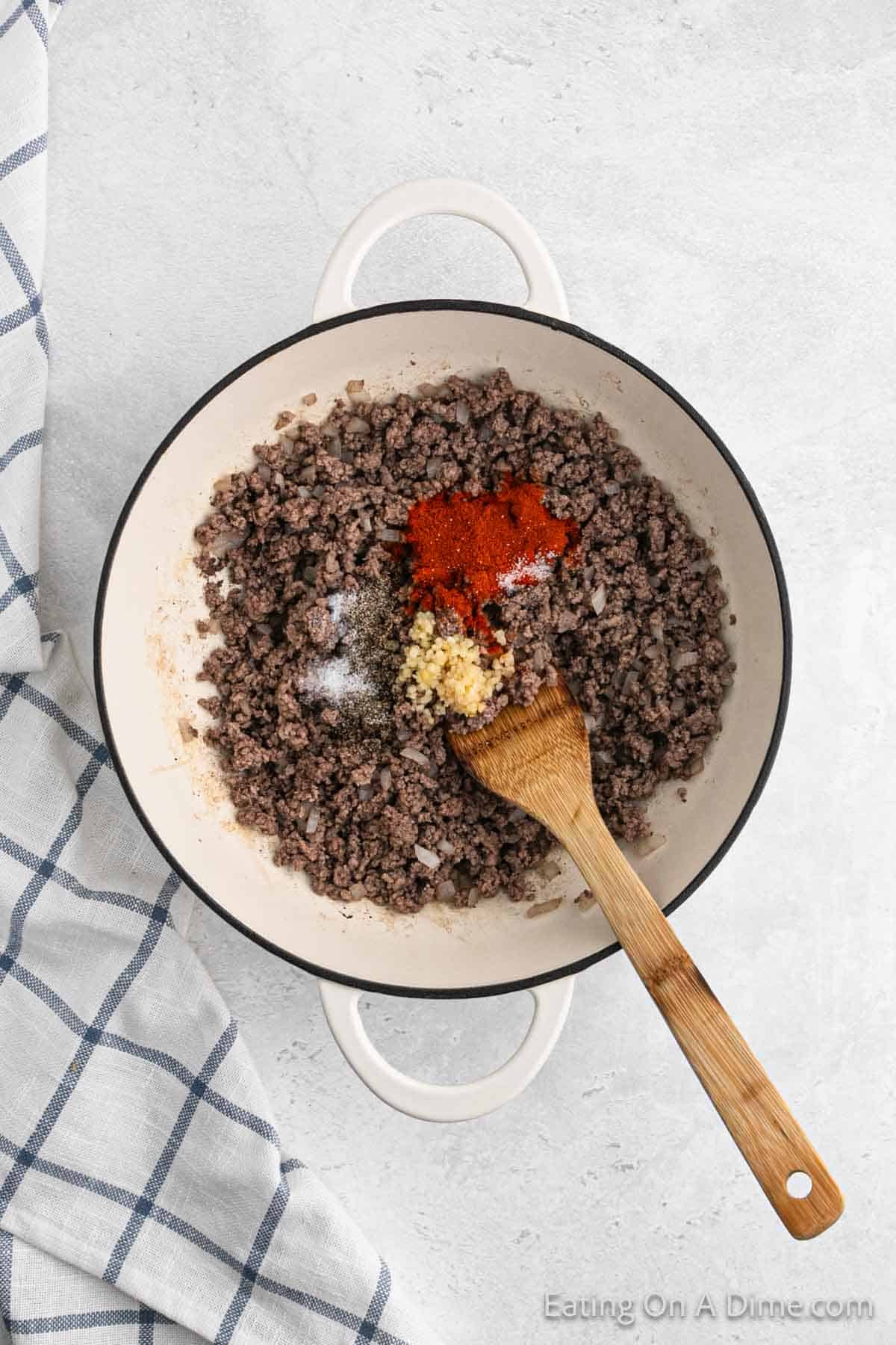A white pot filled with cooked ground beef is being mixed with various spices and minced garlic by a wooden spoon, creating a delectable Homemade Cheeseburger Helper. The pot is sitting on a light gray surface next to a blue-checkered cloth.