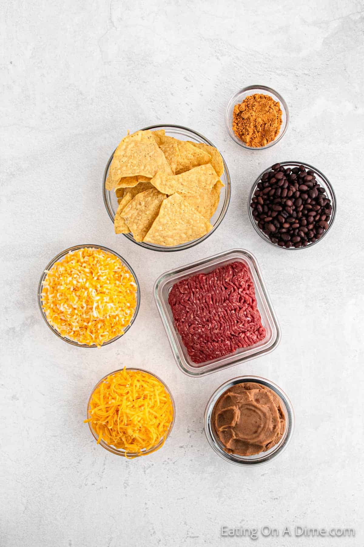 An assortment of taco ingredients is displayed in glass bowls on a textured white surface. Clockwise from top left: tortilla chips, taco seasoning, black beans, refried beans, shredded cheddar cheese, a shredded cheese mixture, and raw ground beef—perfect for crafting oven nachos.