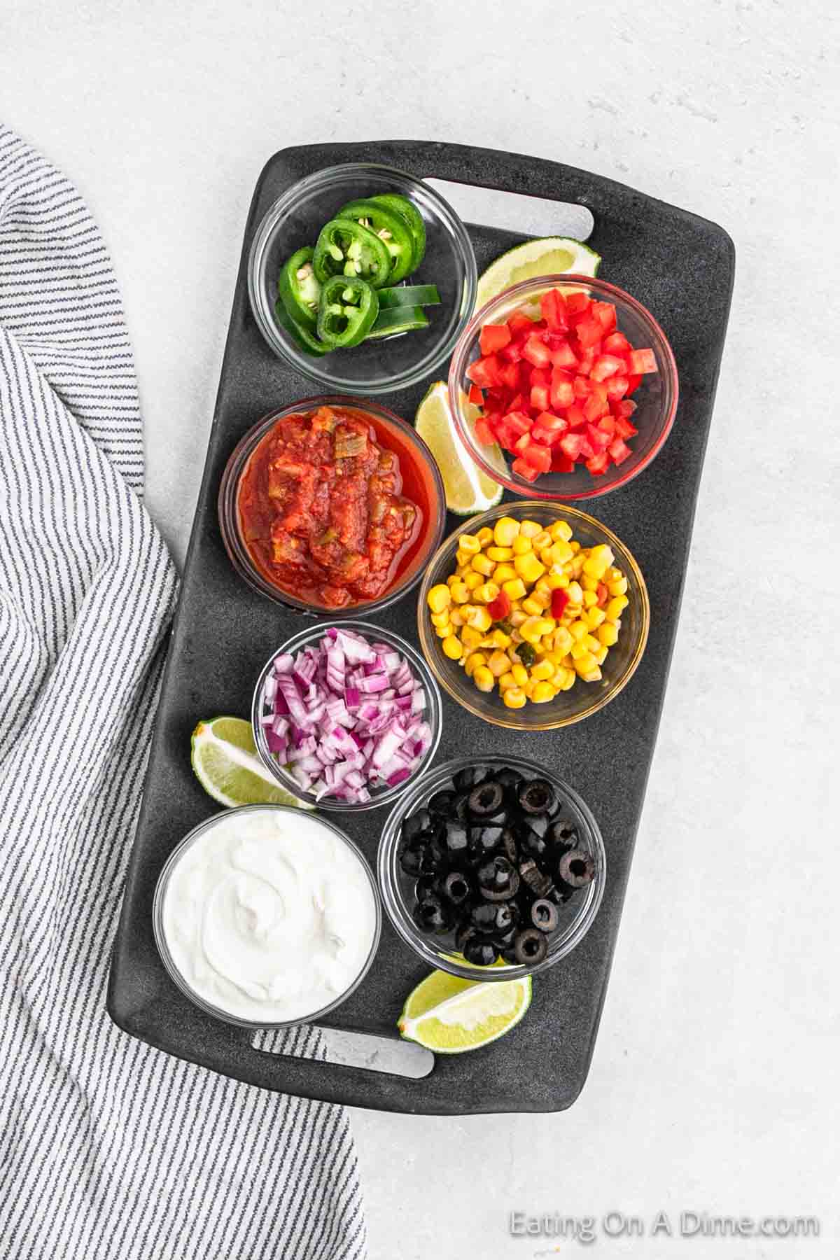 A black serving tray showcases oven nachos with small bowls of sliced jalapeños, salsa, diced tomatoes, corn, red onions, black olives, and sour cream. Lime wedges sit beside the bowls. A striped napkin is partially visible to the left of the tray.