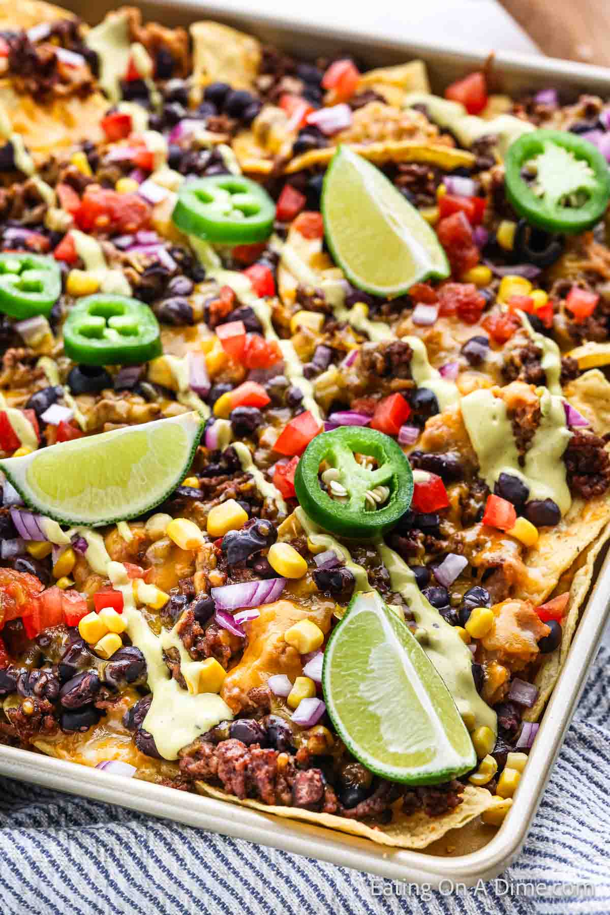 A close-up shot of loaded oven nachos on a tray. The nachos are topped with melted cheese, black beans, ground meat, corn, diced tomatoes, chopped red onions, sliced jalapeños, and avocado cream. There are also lime wedges placed on top of the nachos.