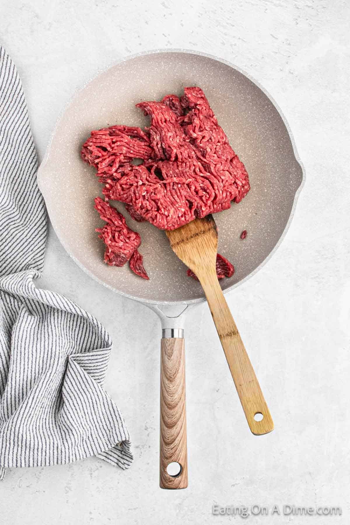 A frying pan with raw ground beef being stirred by a wooden spatula. A striped kitchen towel lies beside the pan on a light-colored countertop. Text "Eating On A Dime.com" is visible in the bottom right corner, perfect for your next batch of oven nachos.