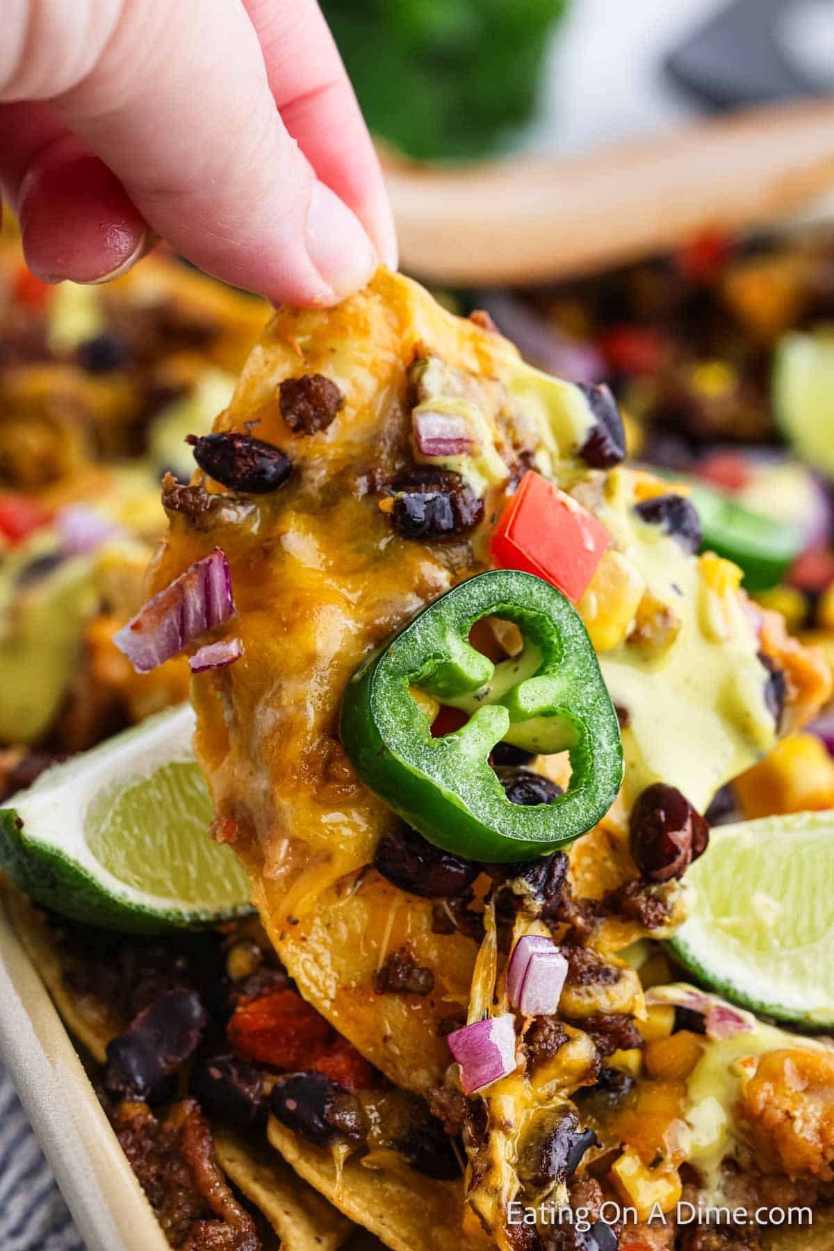 A close-up of a hand holding an oven nacho chip topped with melted cheese, black beans, corn, diced tomatoes, red onions, a slice of jalapeño, and a drizzle of cheese sauce. Lime wedges and additional nachos are visible in the background.