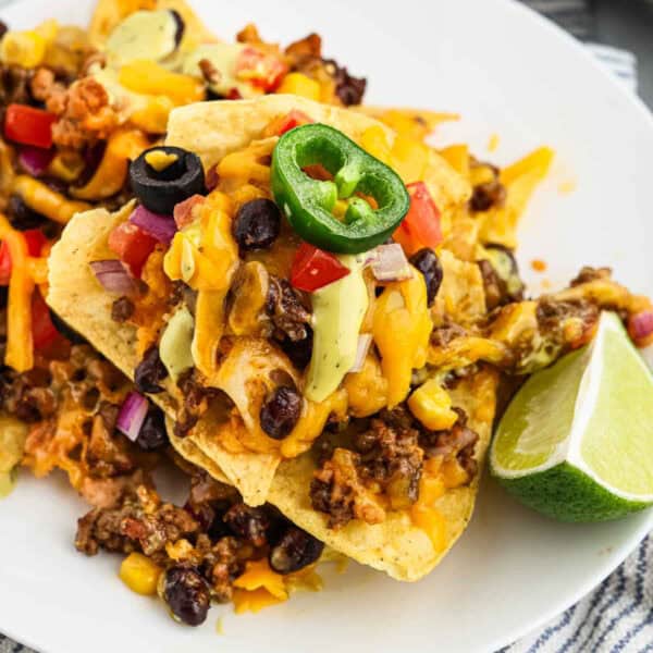 A plate of oven nachos topped with melted cheese, ground beef, black beans, diced tomatoes, red onions, sliced black olives, jalapeño slices, and avocado chunks. A lime wedge is on the side for garnish.