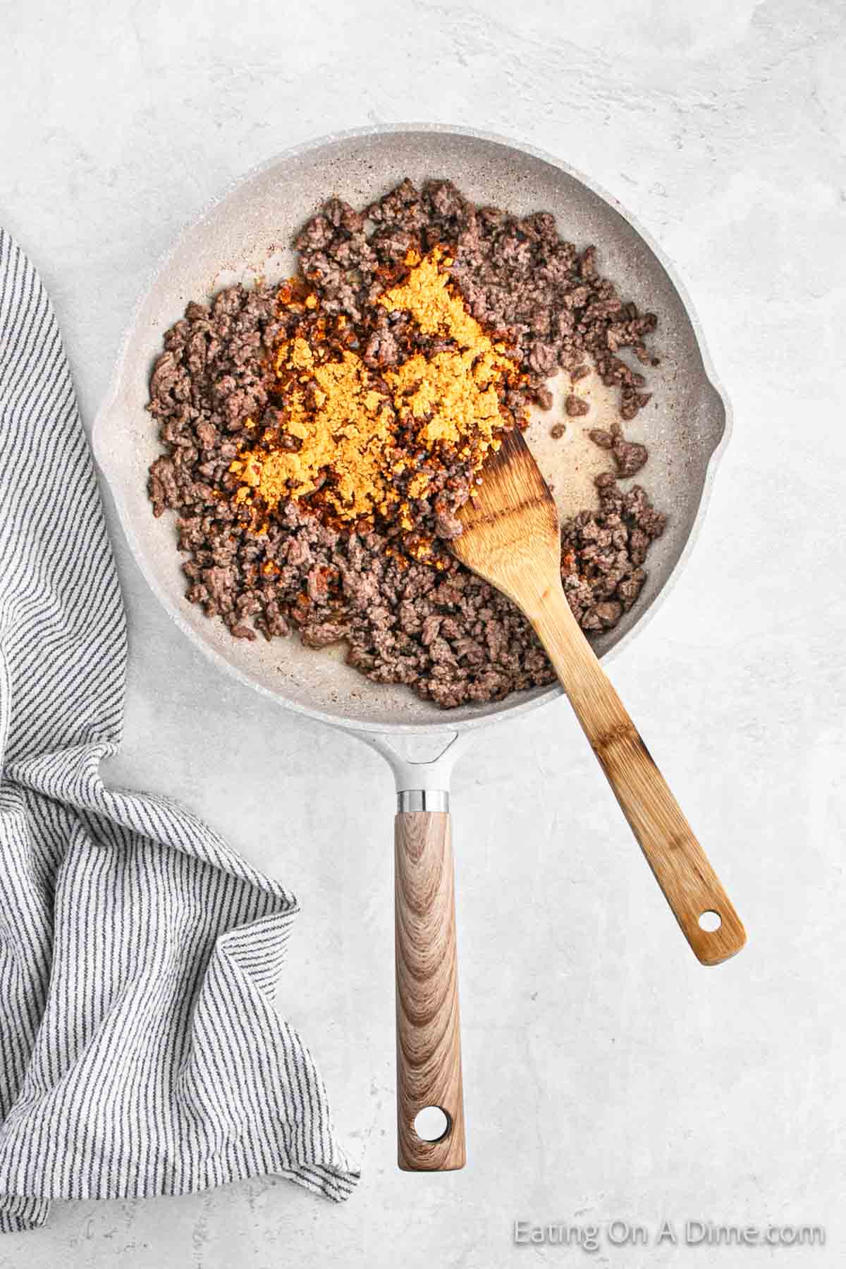 A frying pan containing browned ground beef mixed with taco seasoning sits on a light gray countertop, ready for oven nachos. A wooden spatula rests in the pan, while a gray and white striped kitchen towel is placed next to it. The text "Eating On A Dime.com" is visible in the corner.