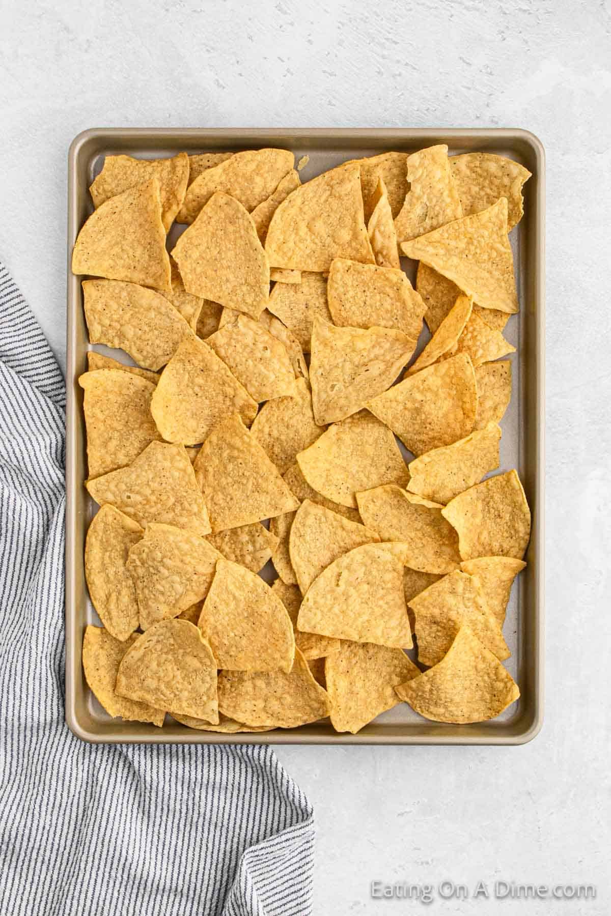 A baking sheet filled with a single layer of triangular corn tortilla chips, perfect for making oven nachos, is placed on a light-colored surface. A gray and white striped cloth is partially visible on the bottom left corner of the image.