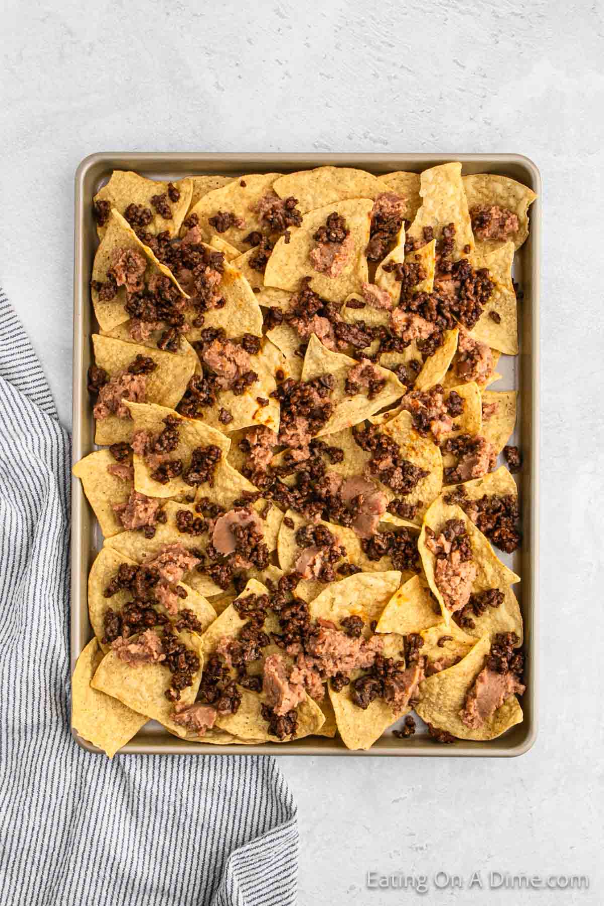 A baking tray filled with oven nachos, topped with melted cheese, ground beef, and beans. A striped kitchen towel is partially visible on the left side of the tray. The tray is on a light gray surface.