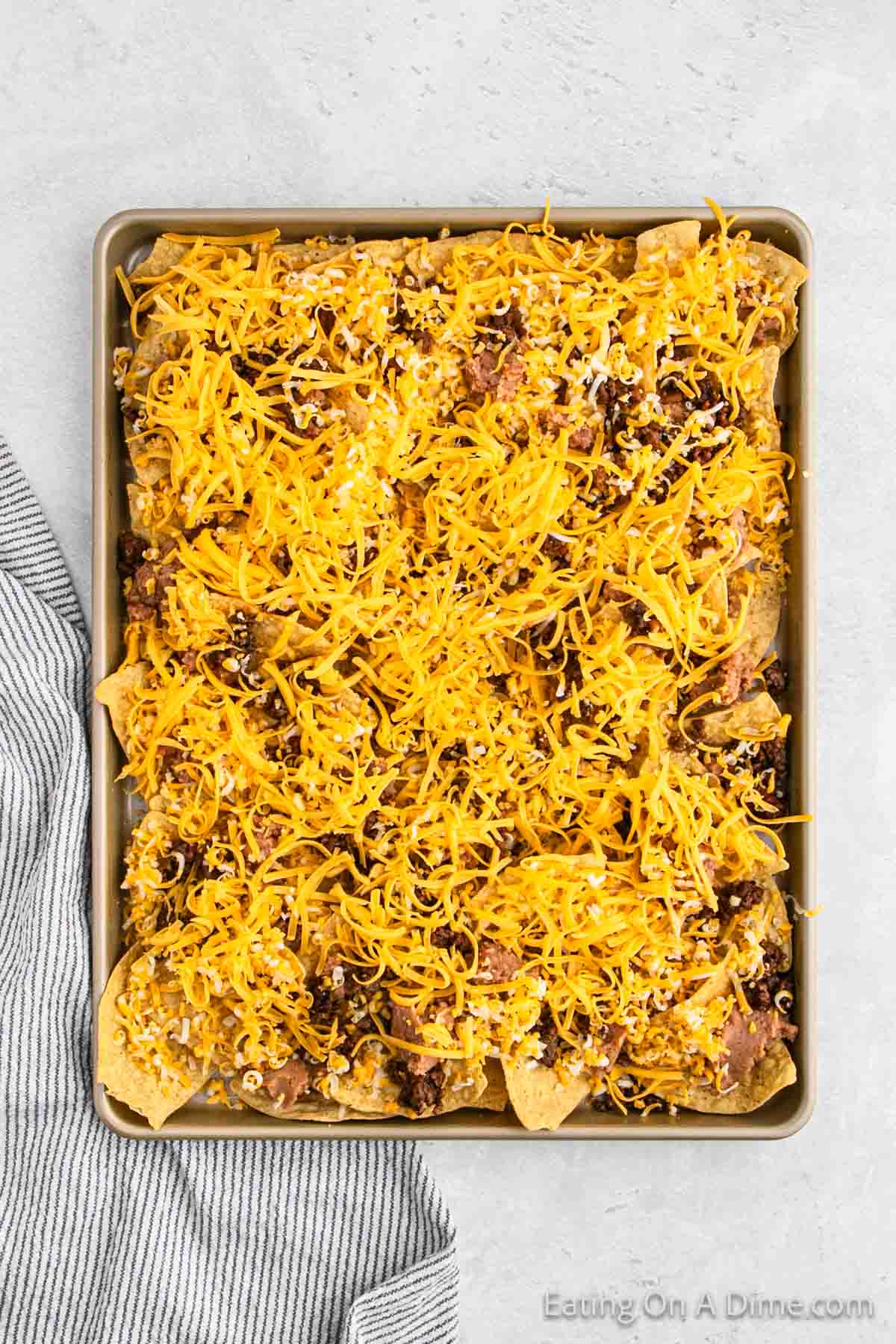 A baking sheet filled with oven nachos, topped with a generous amount of shredded cheese, ground meat, and beans. The sheet is placed on a light-colored surface next to a folded gray and white striped kitchen towel. The website "Eating On A Dime" is visible in the bottom right corner.