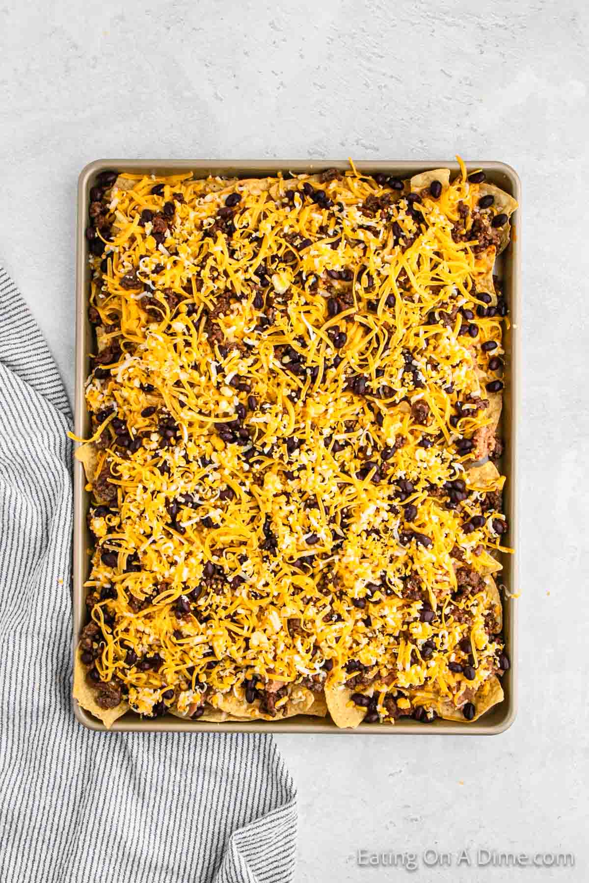 A baking tray filled with oven nachos, topped with shredded cheese, ground beef, and black beans. A gray and white striped cloth is partially visible in the bottom left corner of the image. The background is a light gray surface.