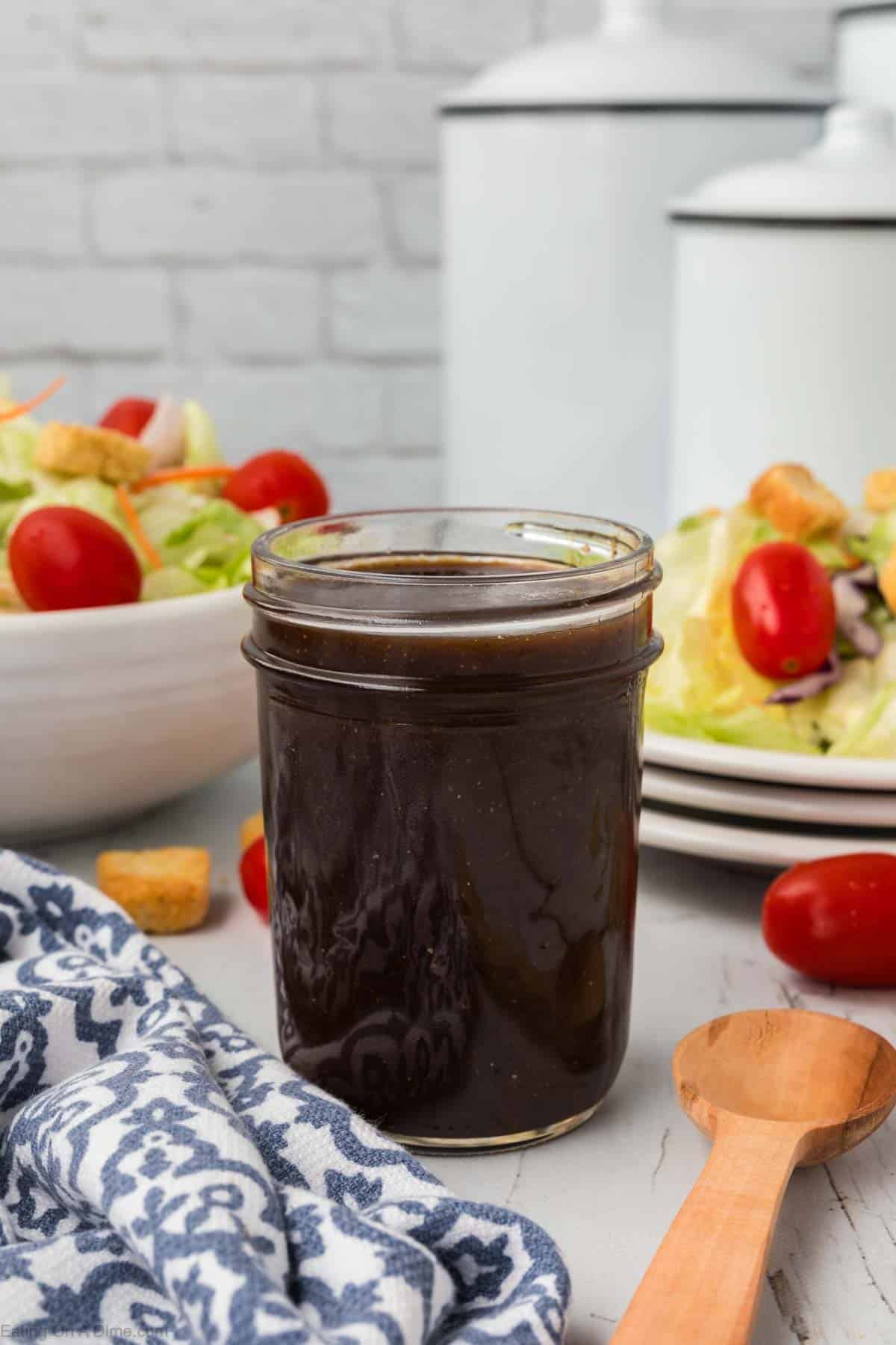 Balsamic Vinaigrette dressing in a mason jar with green salad with cherry tomatoes on a plate and a bowl in the background