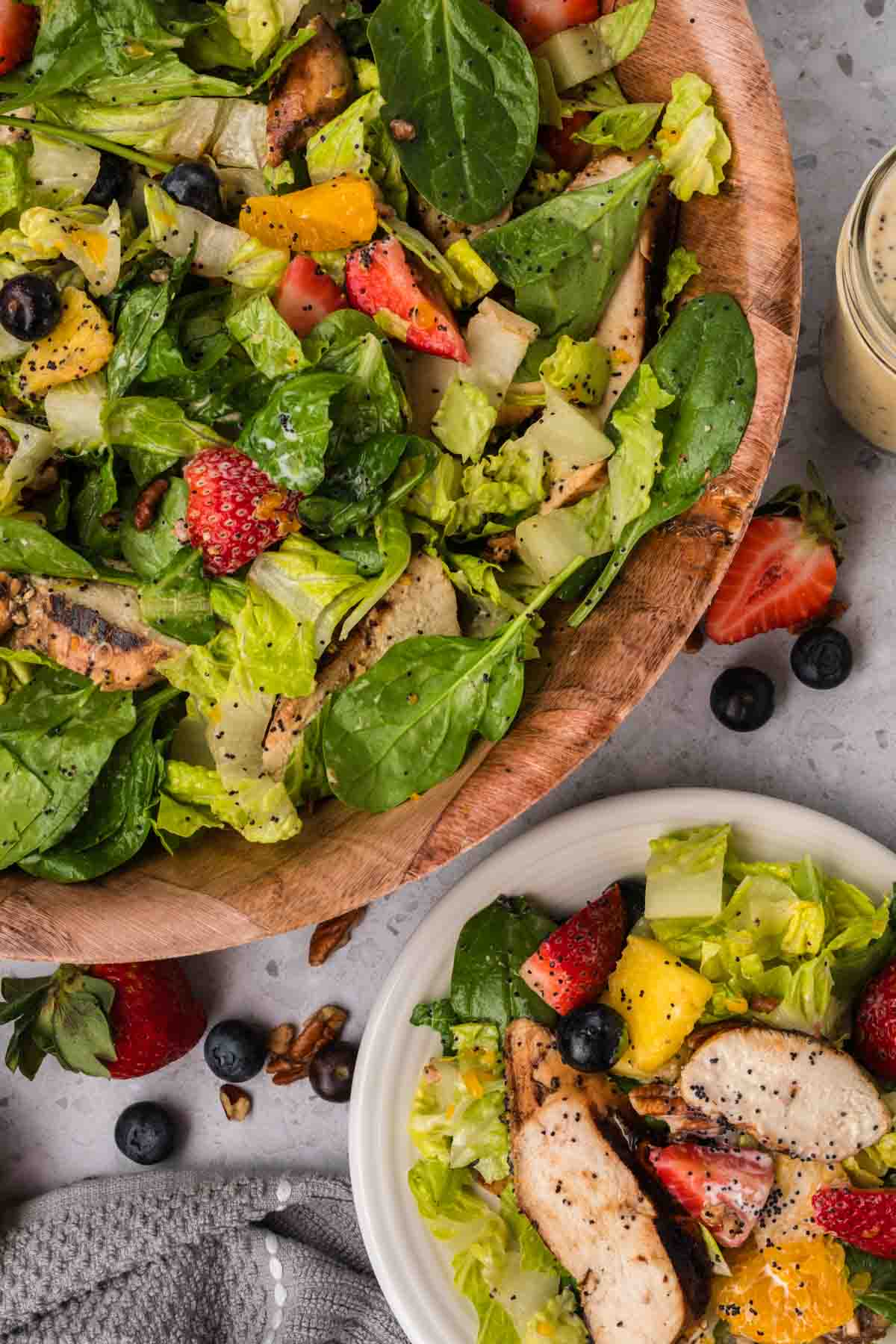 Strawberry Poppyseed Salad in a large bowl topped with slices of grilled chicken, slice strawberries, blueberries and oranges with a plate on the side with strawberry salad