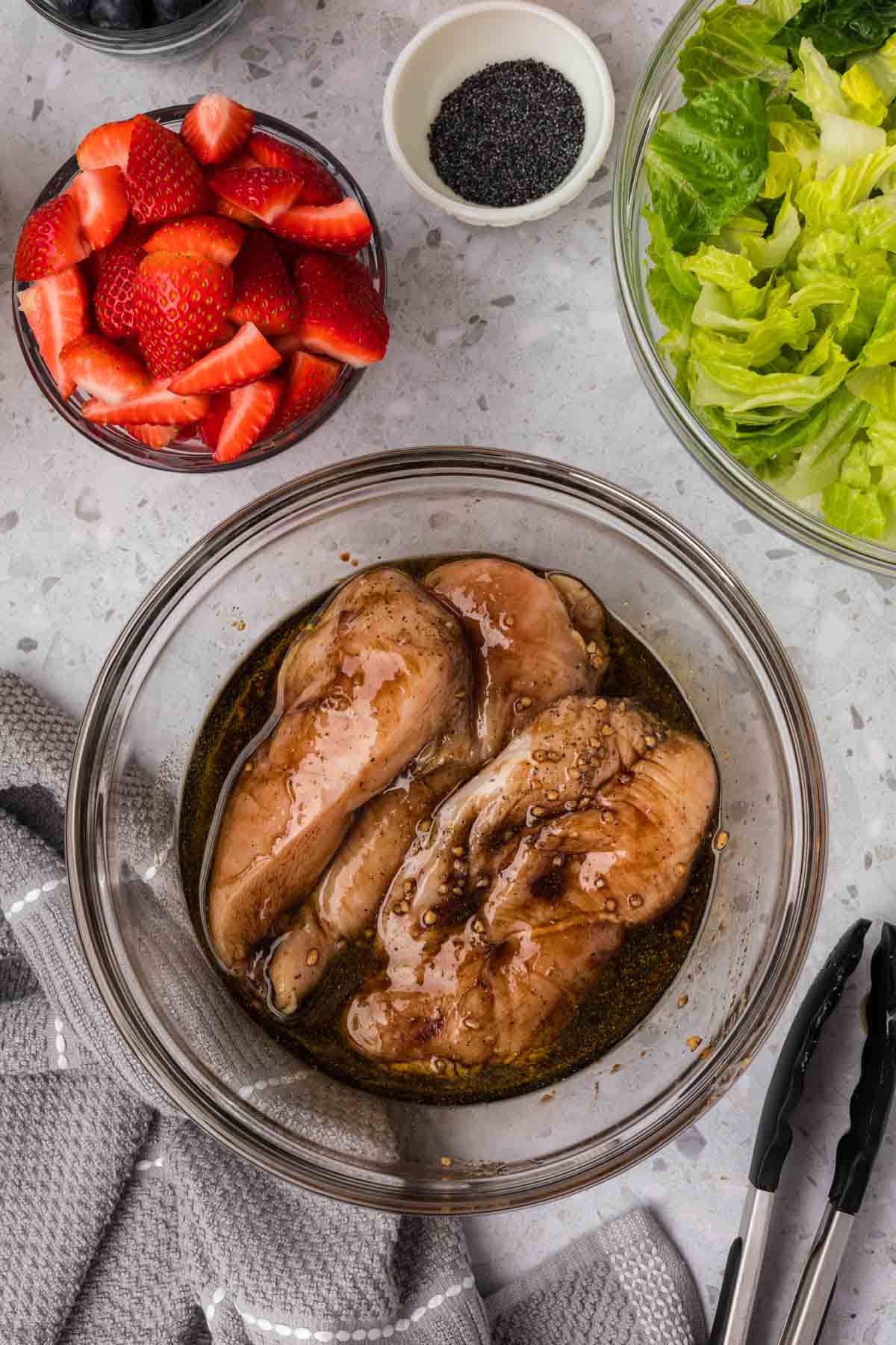 Marinating chicken in a bowl with a side of slice strawberries in a bowl with a bowl of poppyseeds and bowl of chopped romain lettuce