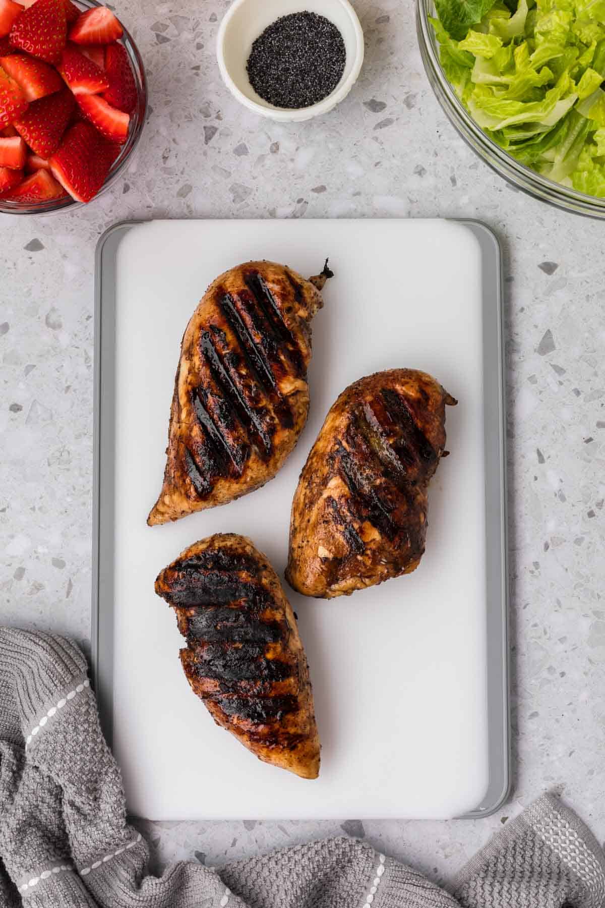 Grilled chicken on cutting board with a bowls of slice strawberries, poppyseed, and chopped romaine lettuce