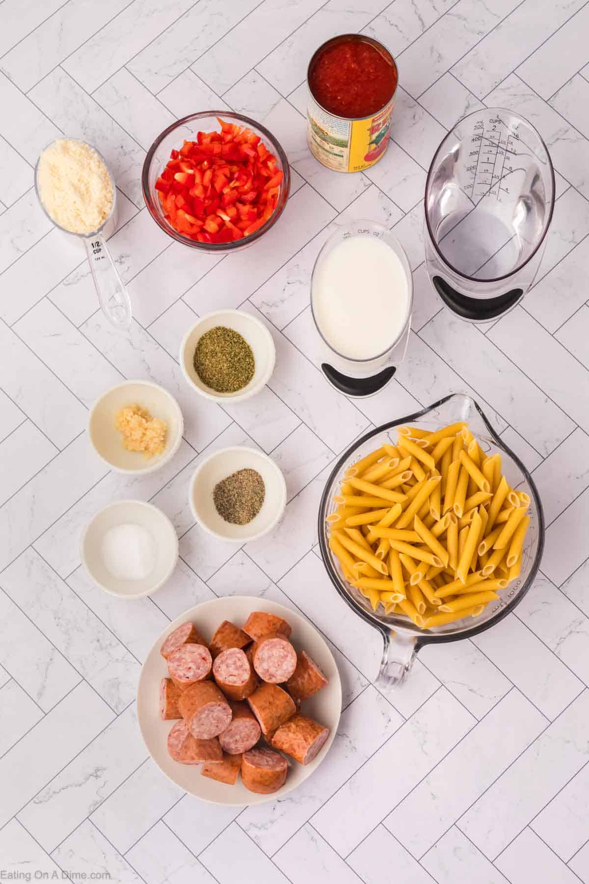 Ingredients for an easy skillet pasta and sausage recipe arranged on a white tiled surface. Items include a measuring cup of milk, a can of tomato sauce, chopped red bell peppers, a cup of grated cheese, seasonings, sliced sausage, minced garlic, and a measuring cup of uncooked penne pasta.