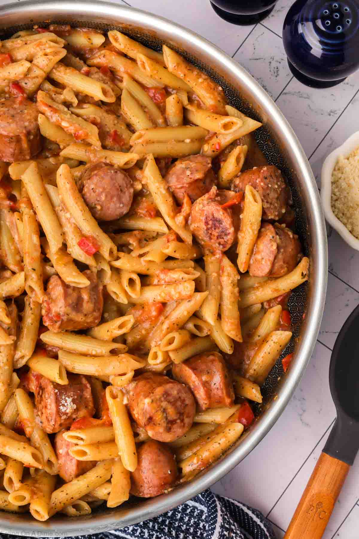 A skillet filled with penne pasta mixed with sliced sausage and diced red bell peppers, coated in a creamy sauce. This easy skillet pasta and sausage recipe rests on a tile surface beside a black kitchen spatula and a small white bowl of grated cheese nearby.