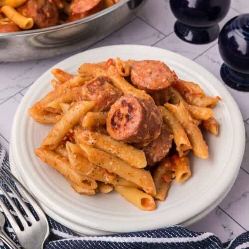 A plate of penne pasta mixed with slices of sausage in a creamy tomato sauce. The easy skillet pasta and sausage recipe is served on a white plate with a fork and knife beside it, and a pan with more pasta is visible in the background. A striped napkin and salt shakers are also present.