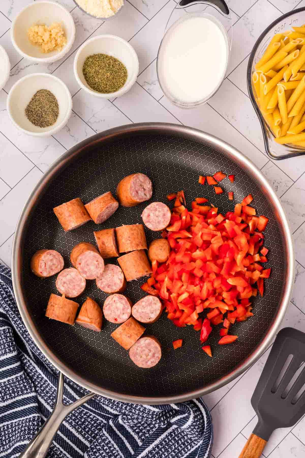 A cooking pan on a stovetop contains sliced sausage and chopped red bell peppers, showing the beginnings of an easy skillet pasta and sausage recipe. Surrounding the pan are small bowls with various ingredients, including dried herbs, milk, grated cheese, and uncooked penne pasta. A striped kitchen towel and a spatula are nearby.