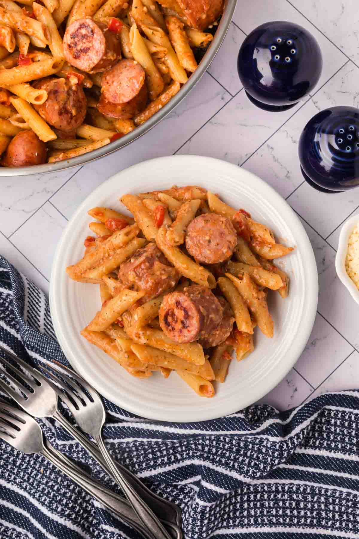 A plate of penne pasta mixed with sliced sausage and vegetables, showcasing an easy skillet pasta and sausage recipe, served on a white plate. In the background, a pot with more pasta sits beside a striped cloth, a few forks, and two black pepper and salt shakers. The table's light tiles complete the scene.