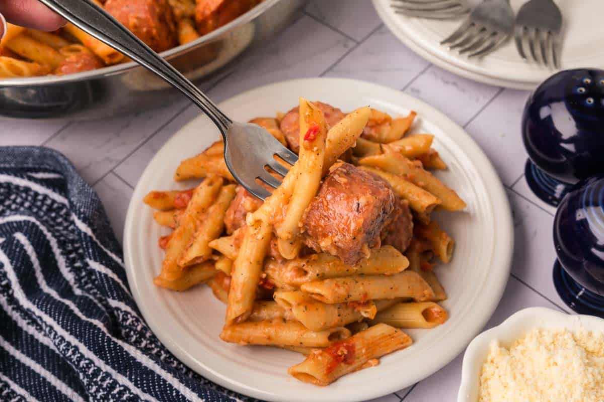 A fork lifting a bite of penne pasta with a meatball from a plate. The pasta is covered in a tomato-based sauce. In the background, there is a striped napkin, reminding you of an easy skillet pasta and sausage recipe, along with a pan of more pasta, plates, and bowl of grated cheese. Salt and pepper shakers are also visible.