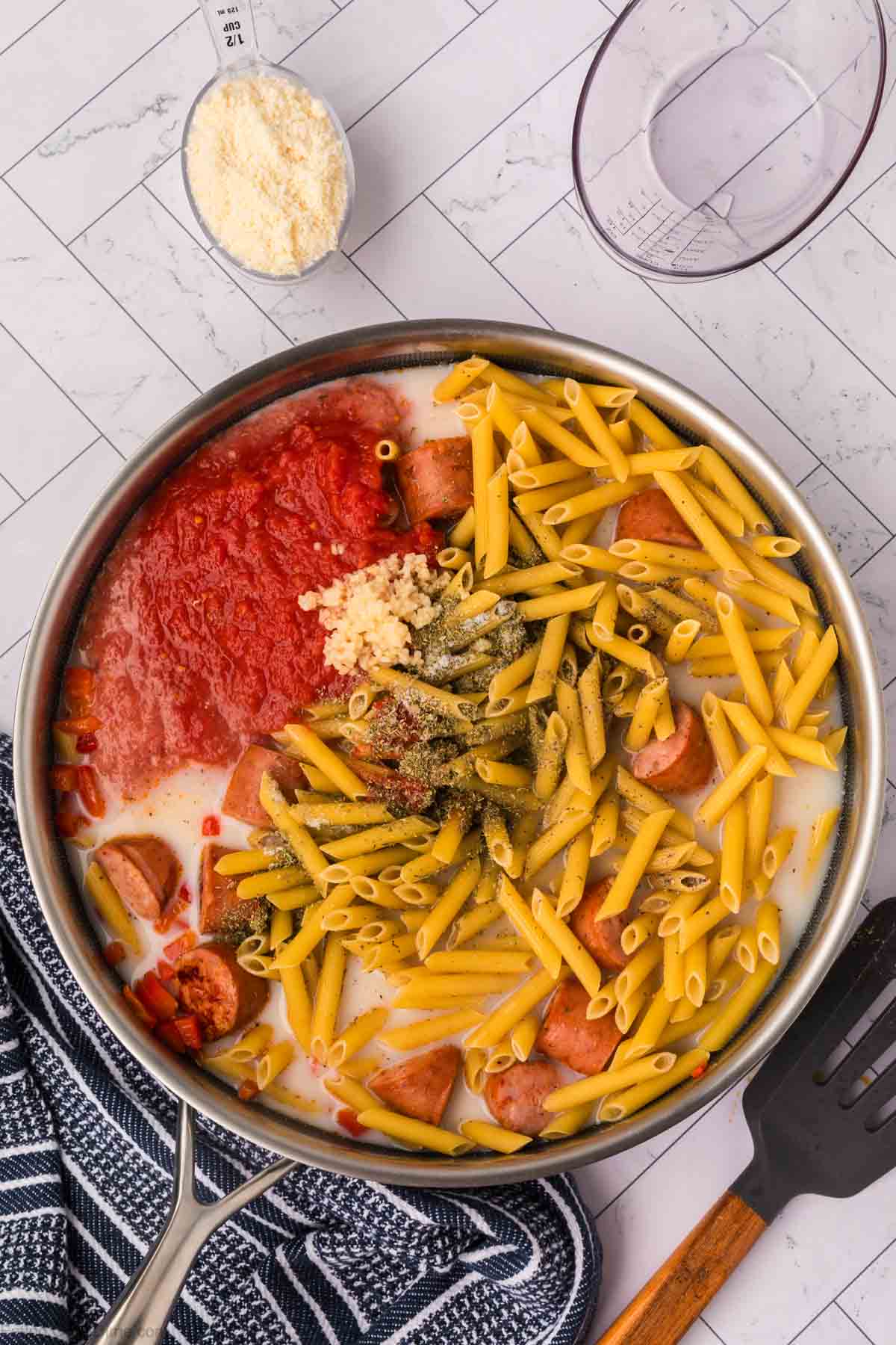 A stainless steel pan filled with uncooked penne pasta, raw sausage slices, tomato sauce, minced garlic, and various seasonings all mingling with milk for an easy skillet pasta and sausage recipe. A black and white dish towel, a measuring spoon with grated cheese, and a clear measuring cup are nearby.