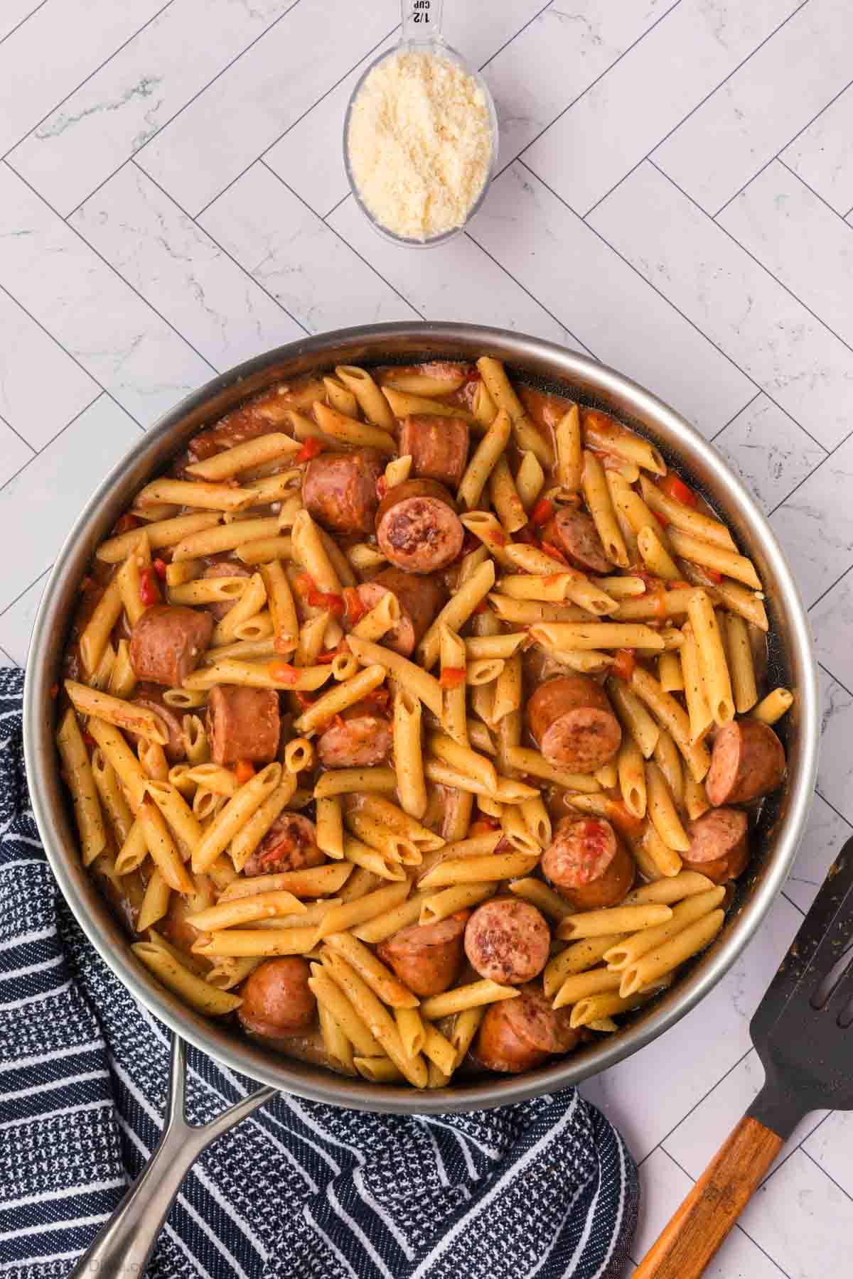 A skillet filled with penne pasta mixed with sliced sausage and red peppers sits on a tiled surface next to a measuring cup with grated cheese. An easy skillet pasta and sausage recipe, it rests beside a dark-striped kitchen towel, with a spatula partially visible.