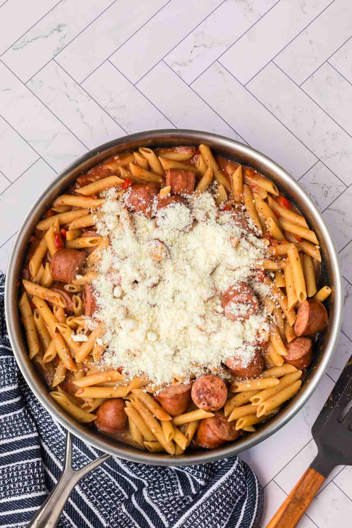 A skillet filled with penne pasta, slices of sausage, and red bell peppers, all topped with a generous layer of grated Parmesan cheese. This easy skillet pasta and sausage recipe rests on a white, tile-patterned surface beside a dark-striped kitchen towel and a spatula.