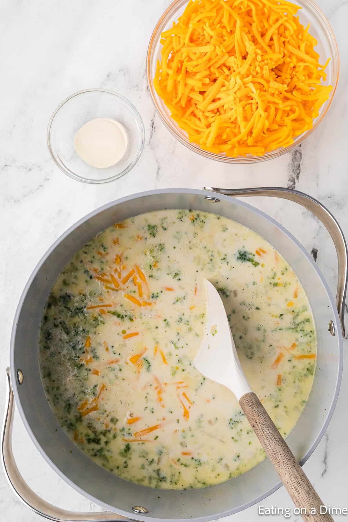 A pot of creamy Keto Broccoli Cheese Soup, with bits of green vegetables, is being stirred with a white spatula. Beside the pot, there's a bowl filled with grated cheddar cheese and a small dish containing light-colored seasoning. "Eating on a Dime" is printed in the corner.