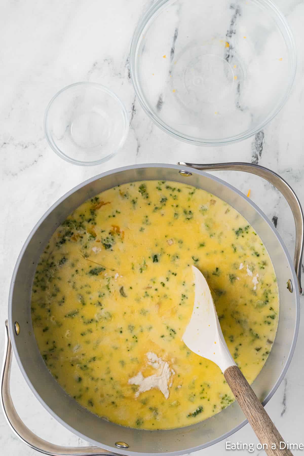 A pot of creamy Keto Broccoli Cheese Soup with visible pieces of broccoli and chunks of meat sits on a marble countertop. A white and wooden spoon rests in the pot, while two empty glass bowls await beside it. The image features the text "Eating on a Dime" in the bottom right corner.