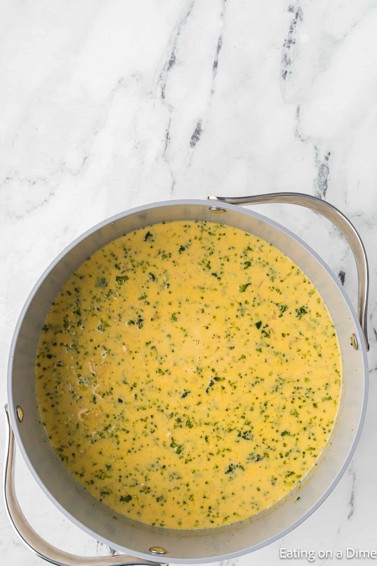 A large silver pot on a white marble countertop contains a creamy, yellow Keto Broccoli Cheese Soup with visible herbs and small green vegetable pieces. The soup appears to be thick and well-blended. The text "Eating on a Dime" is partially visible in the bottom right corner.