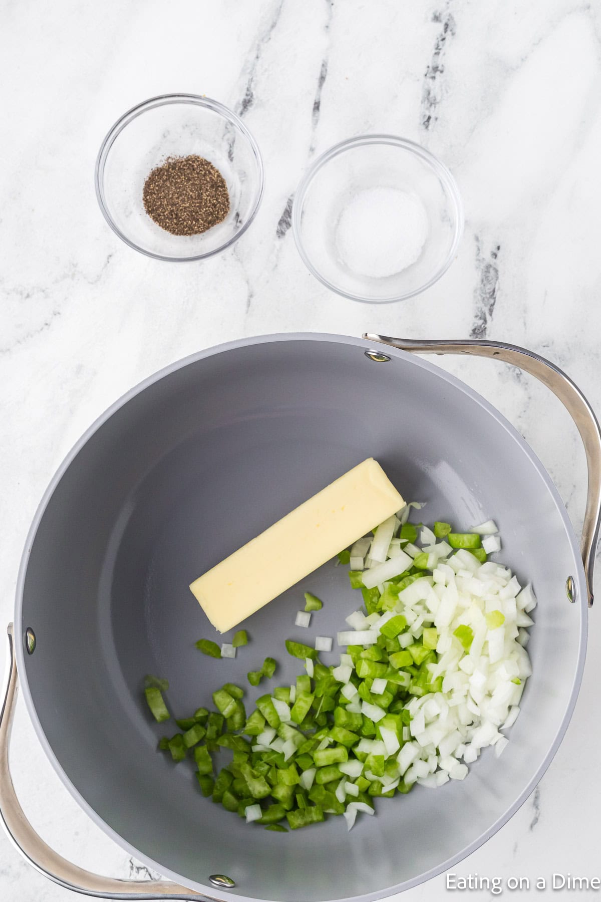 A grey pot on a marble countertop contains chopped onions and green bell peppers with a stick of butter, ready to start the Keto Broccoli Cheese Soup. Two small glass bowls nearby hold black pepper and salt. Text at the bottom right corner reads "Eating on a Dime.