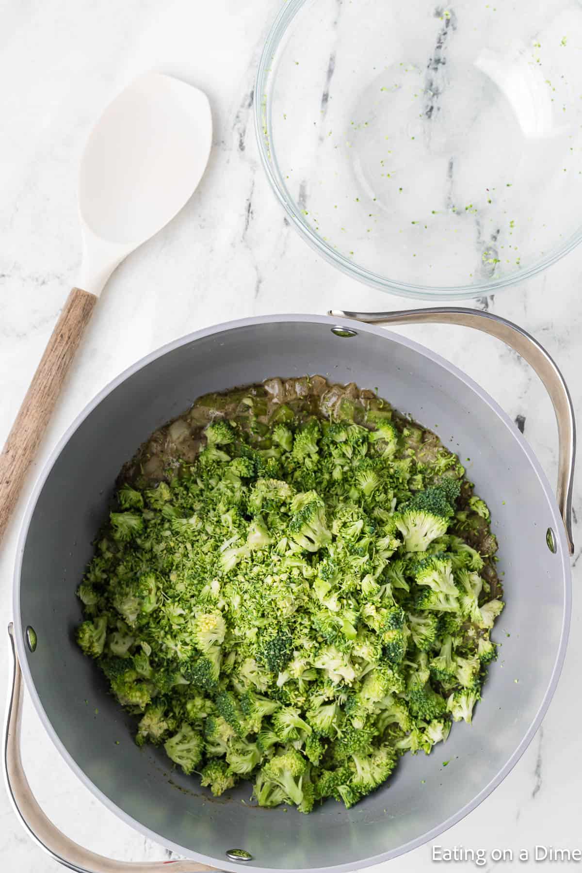 A pot on a marble countertop filled with chopped broccoli and sautéed onions, ready to be transformed into a Keto Broccoli Cheese Soup. A large white spoon with a wooden handle is placed beside the pot. An empty glass mixing bowl nearby holds some chopped broccoli remnants inside.