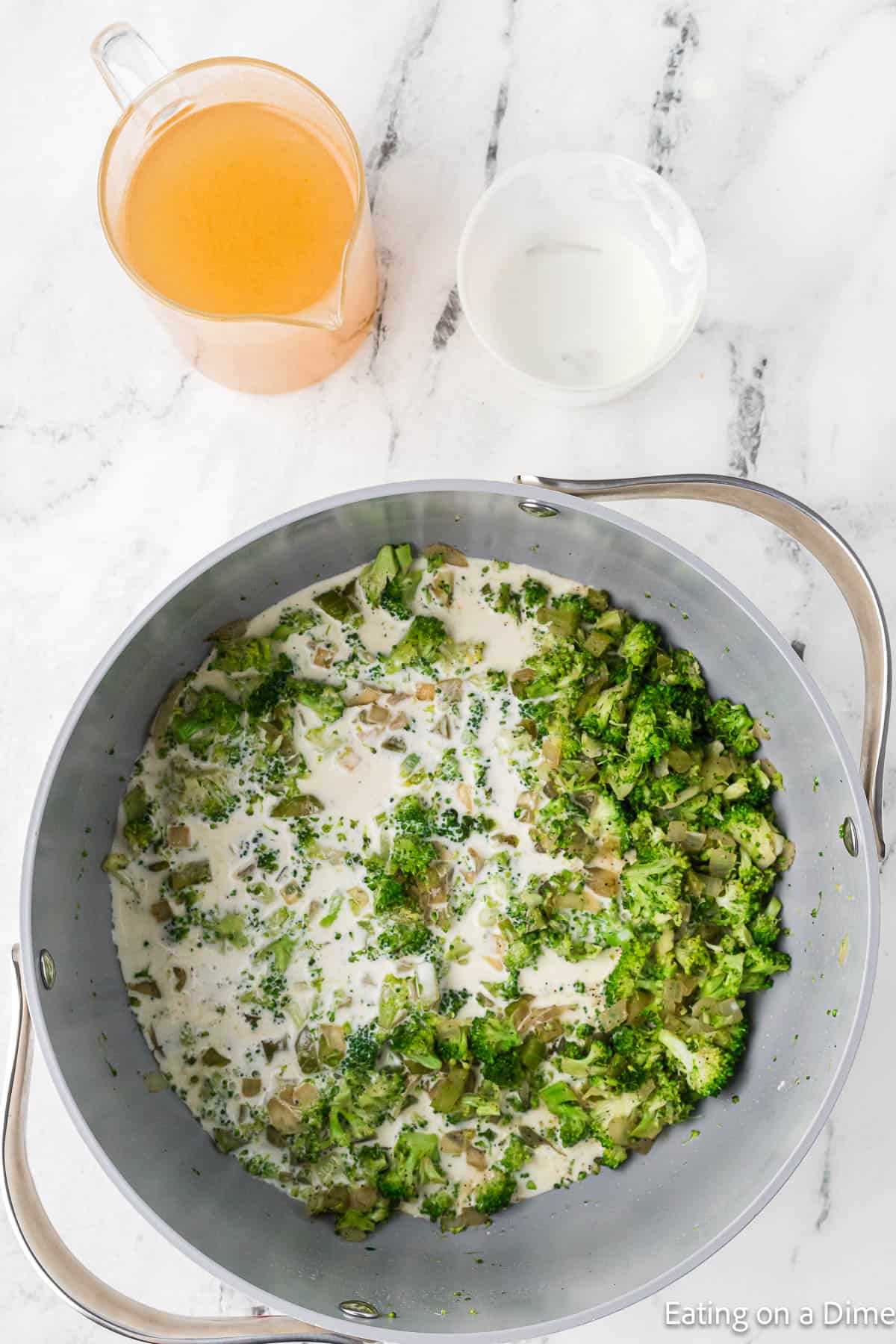 A pot on a marble countertop contains broccoli florets and a creamy mixture, perfect for Keto Broccoli Cheese Soup. Beside the pot, there is a clear pitcher with a yellow liquid and a small white bowl. The picture captures the ingredients being prepared for this delicious recipe.