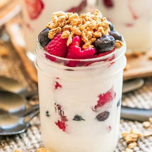 A glass jar filled with creamy yogurt, topped with fresh blueberries, raspberries, and a generous layer of crunchy granola, making for an easy fruit and yogurt parfait. A few more spoons and jars with similar contents are visible in the blurred background.