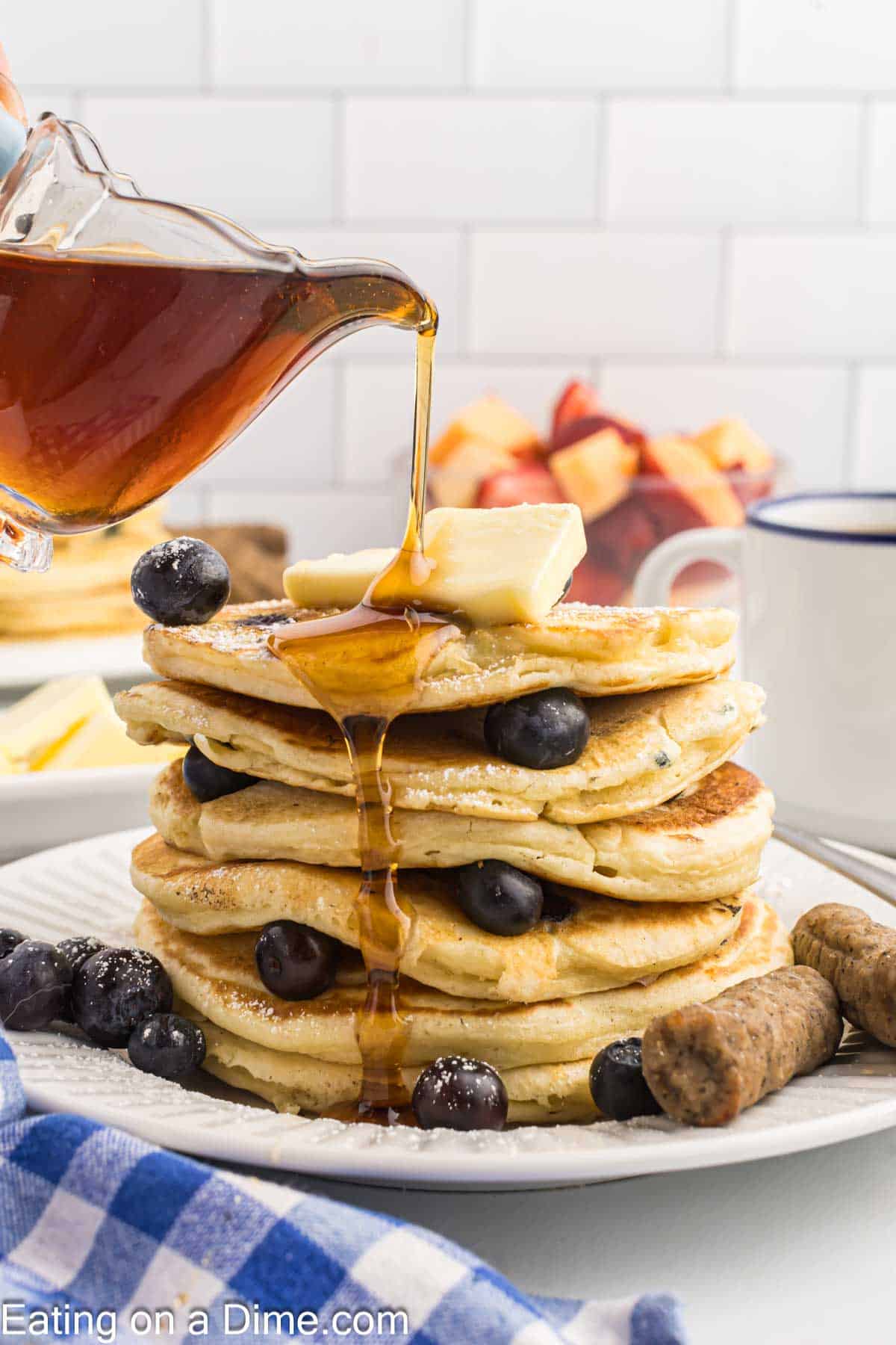 Syrup is being drizzled over the top of pancakes stacked on a plate with fresh blueberries