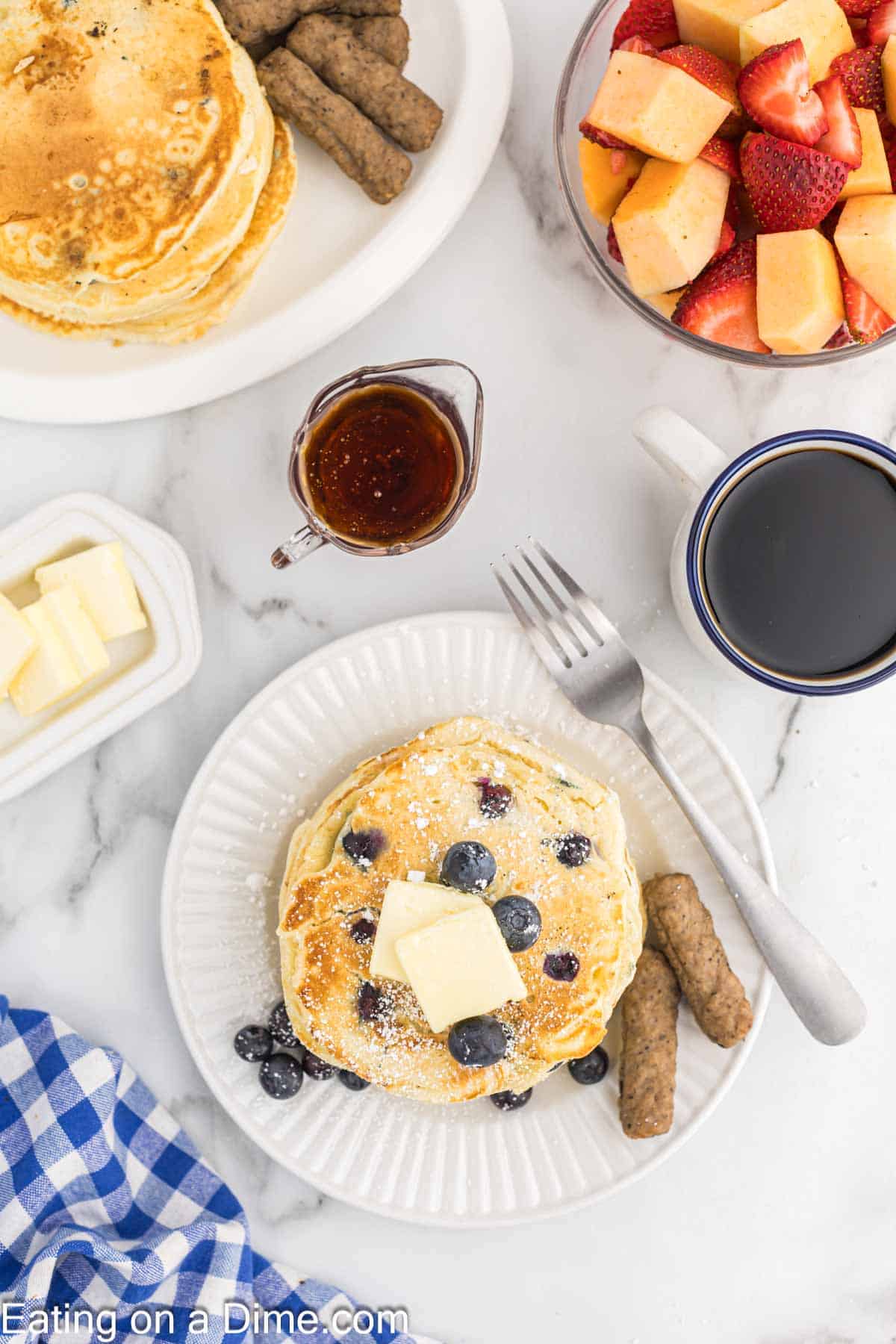 Blueberry pancakes stacked on a plate topped with butter, with a side of sausage links. A bowl of syrup, a cup of coffee and a bowl of fresh fruit is on the side
