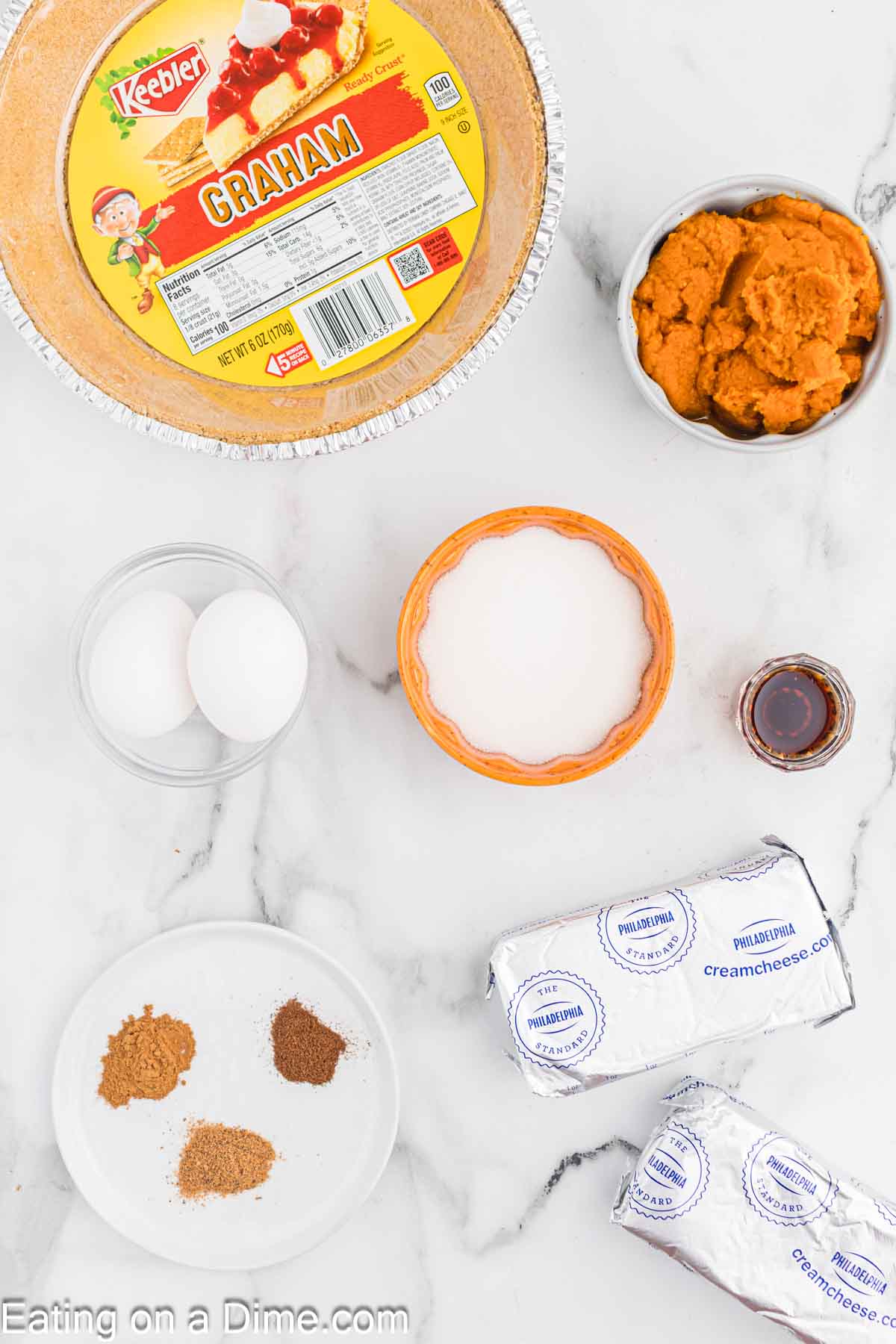 Top view of various ingredients for making a pumpkin cheesecake on a marble surface. Visible are a Keebler graham pie crust, a bowl of pumpkin puree, two eggs, a bowl of sugar, a plate with spices, two packs of cream cheese, and a small container of vanilla extract.