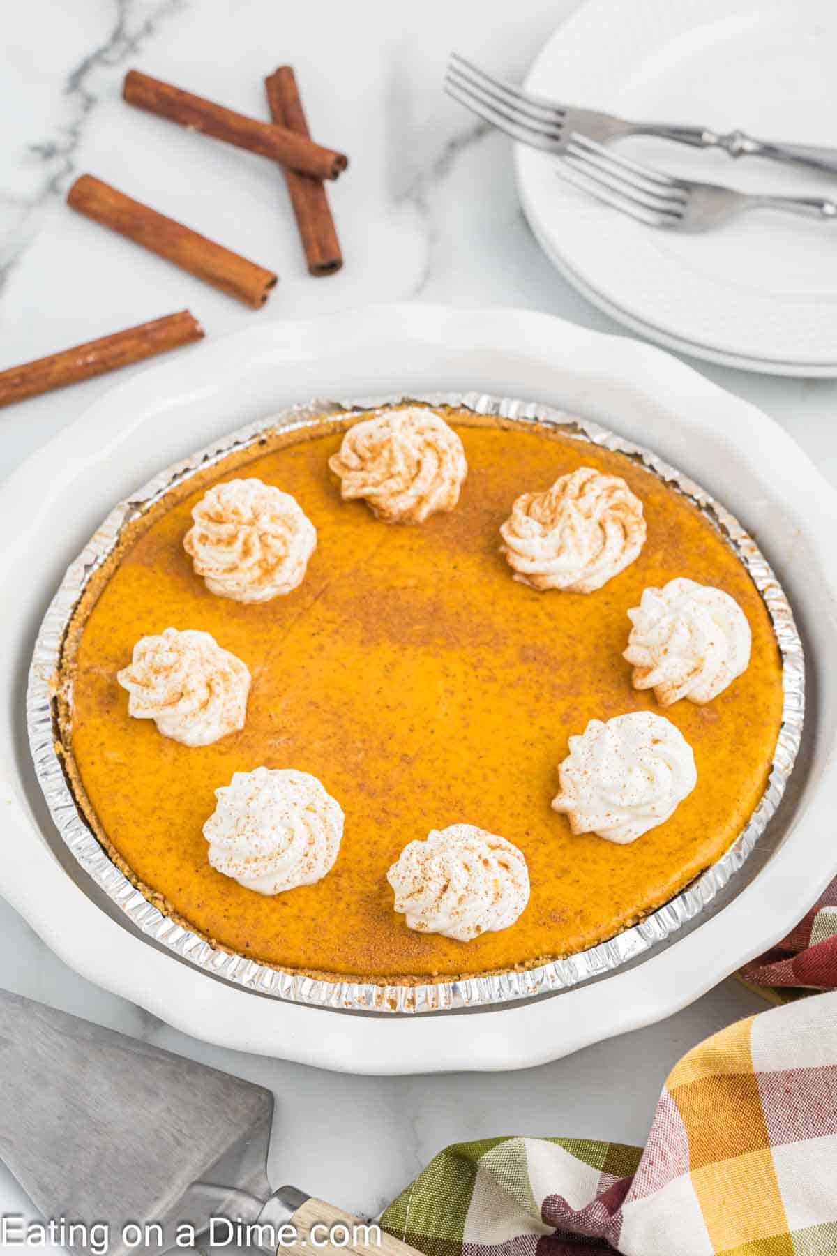 A whole pumpkin pie in a foil pie tin is topped with dollops of whipped cream evenly spaced around the edge. Three cinnamon sticks and two stacked white plates with forks are in the background. A colorful fabric napkin is partially visible in the corner, hinting at flavors similar to a delightful pumpkin cheesecake.