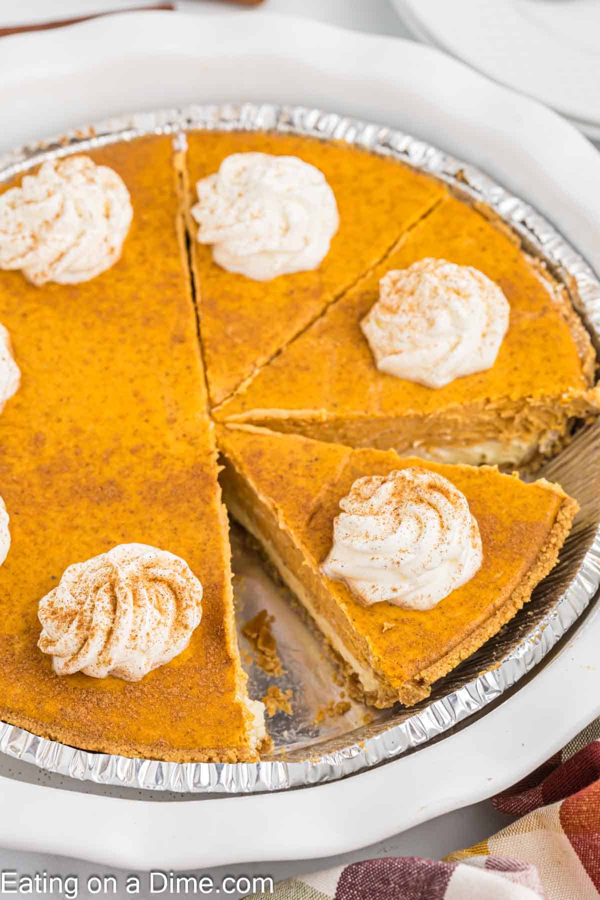 A pumpkin cheesecake pie in a silver baking dish, topped with swirls of whipped cream and sprinkled with cinnamon. A slice has been removed, revealing its creamy interior. The pie is placed on a white surface, with a seasonal cloth partially visible at the bottom.
